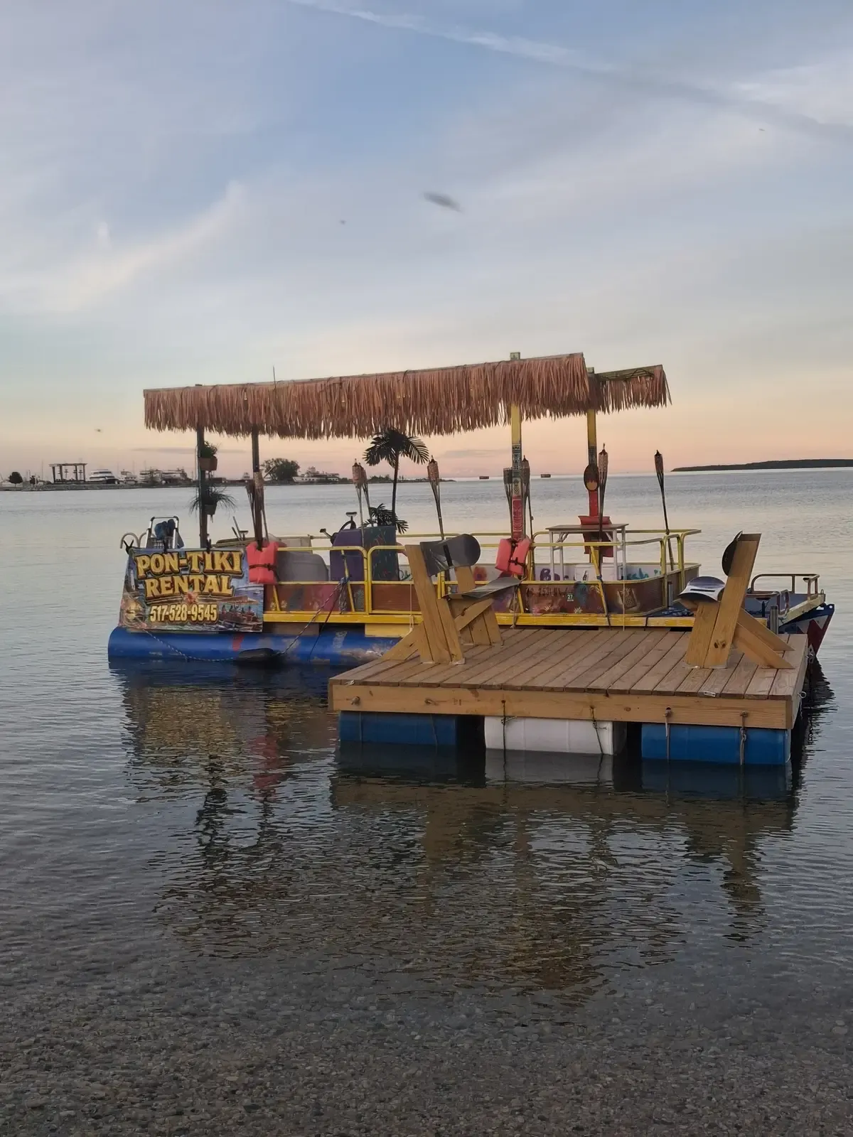 The custom Pon Tiki pontoon boat docked at sunset with tiki torches along the shore