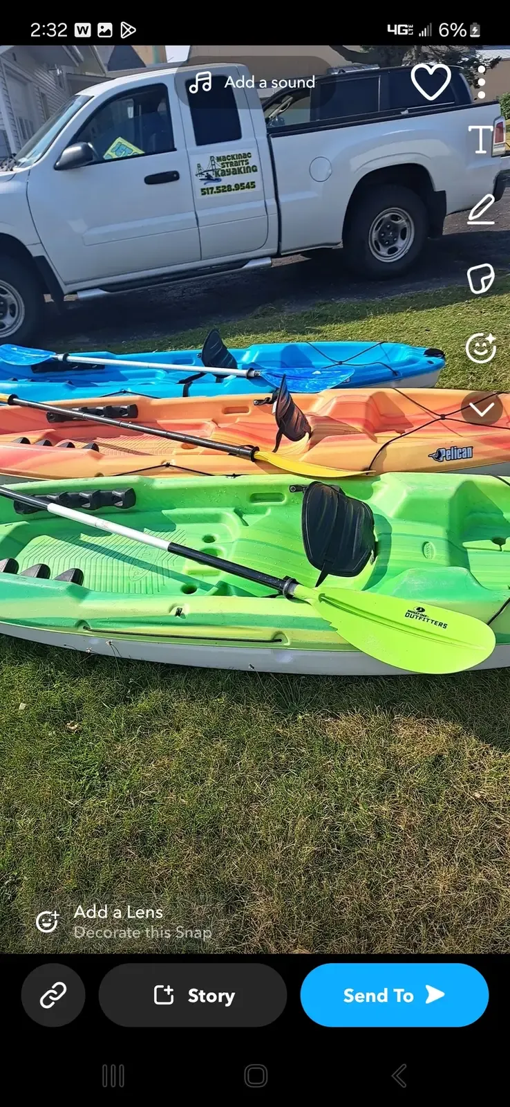 A row of colorful rental kayaks with paddles lined up on the grass beside the Mackinac Straits Kayaking truck